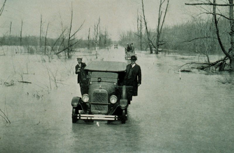 A classic black and white photo that dramatically shows the effects of the great Mississippi flood in 1927. Two men lean out the door and stand outside as the cameraman takes the photo.